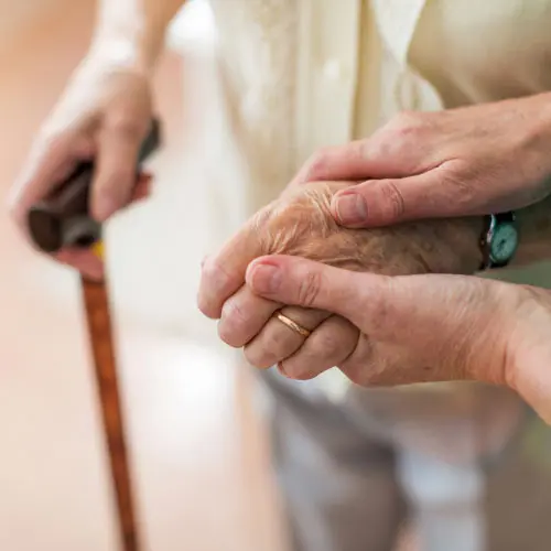 Caregiver holding an older adult's hands while using a cane, representing ongoing long-term care (LTC) training, elder care, mobility assistance, and compassionate caregiving.