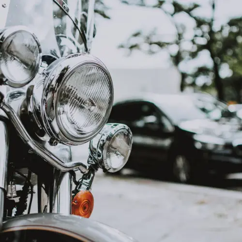 Close-up of chrome motorcycle headlights with a parked car blurred in the background, representing advanced thinking about the personal auto policy
