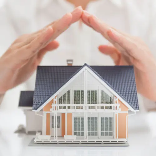 Hands forming a protective roof shape over a small model house, representing a property and casualty insurance primer