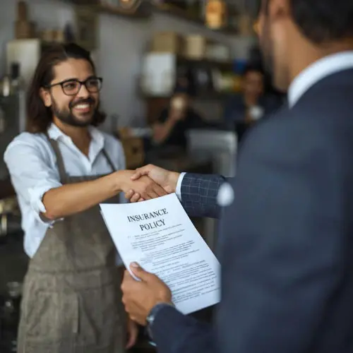 Small business owner in an apron shaking hands with an insurance agent holding an insurance policy document in a shop setting, representing small and home business insurance