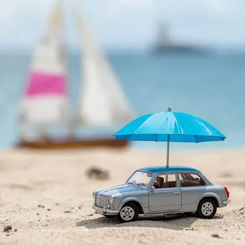 Toy car sheltered by a small blue umbrella on a beach with a sailboat in the background, representing umbrella, watercraft, and automobile insurance