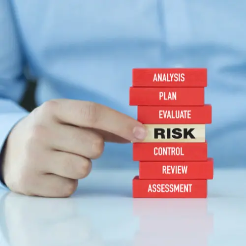 Hand stacking red blocks labeled analysis, plan, evaluate, risk, control, review, and assessment, representing varieties of risk